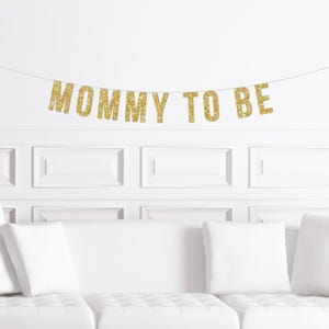 May include: A gold glitter "MOMMY TO BE" banner is suspended above a white sofa with white cushions. The banner is strung across a white wall with rectangular panelling, creating a celebratory atmosphere.