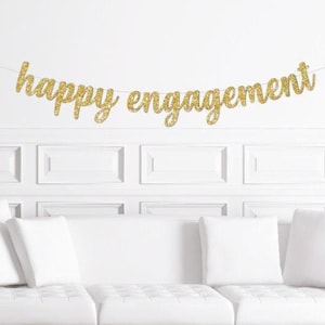 May include: A gold glitter "happy engagement" banner hangs above a white sofa. The banner is strung across a white wall with decorative panelling. The letters are in a cursive font and sparkle.