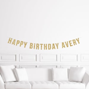 May include: A gold glitter banner that reads "HAPPY BIRTHDAY AVERY" hangs against a white wall. The banner is strung across a thin string. Below the banner is a white couch with white pillows.