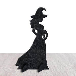 May include: Black silhouette of a pregnant witch wearing a pointed hat and flowing dress. The witch's belly has a cutout of baby footprints. The Halloween decoration is made of a textured material and stands on a wooden surface.