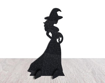 May include: Black silhouette of a pregnant witch wearing a pointed hat and flowing dress. The witch's belly has a cutout of baby footprints. The Halloween decoration is made of a textured material and stands on a wooden surface.