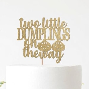 May include: A gold glitter cake topper with the phrase "two little dumplings on the way." The topper features two smiling dumpling illustrations. The topper is placed on top of a white cake.
