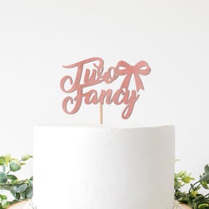 May include: A pink glitter cake topper with the words "Two Fancy" and a bow design. The topper is on a wooden stick, placed on a white cake. Green foliage is at the base of the cake.