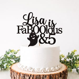 May include: A black glittery cake topper featuring the text Lisa is FaBOOlous & 5 with a ghost graphic. The topper is inserted into a white frosted cake. The text is written in a combination of cursive and block lettering with a smiling ghost.