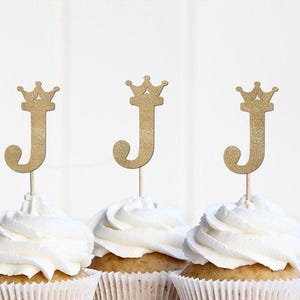 May include: Three cupcakes with white frosting, each topped with a gold glitter letter "J" and a crown. The cupcakes are in white paper liners, perfect for a birthday or celebration.