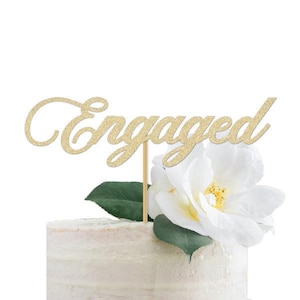 May include: A gold glitter "Engaged" cake topper on a white cake, with a white flower and green leaves. The topper is attached to a wooden stick. The cake has a light-coloured frosting.