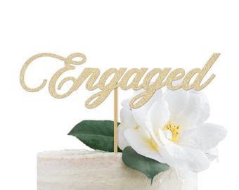 Peut inclure: Un cake topper doré à paillettes avec le mot "Engaged" sur un gâteau blanc, avec une fleur blanche et des feuilles vertes. Le cake topper est fixé à un bâton en bois. Le gâteau a un glaçage clair.