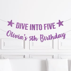 May include: A purple glitter banner with the text "DIVE INTO FIVE" and "Olivia's 5th Birthday" strung across a white wall. Two star-shaped decorations flank the top banner. The banner is a festive decoration for a birthday party.