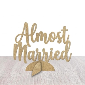 May include: A wooden sign with the words "Almost Married" in a cursive font. The sign is a light tan color and has a stand for display. The background is white.