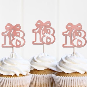 May include: Three cupcakes with white frosting and decorative rose gold number 18 toppers. The toppers have a bow design above the numbers. The cupcakes are in white paper liners.