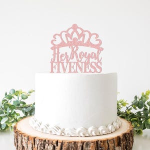 May include: A white iced cake with a pink glitter crown cake topper that reads "Her Royal Fiveness." The cake sits on a wooden cake stand with a decorative white icing border. Greenery surrounds the cake.