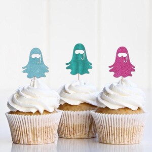 May include: Three cupcakes with white frosting and decorative glitter ghost toppers in teal, green, and pink. The cupcakes are in white paper liners, and the background is a plain white.