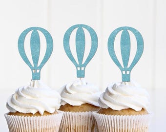 May include: Three cupcakes topped with white frosting and light blue glittery hot air balloon toppers. The cupcakes are in white paper liners, and the background is a plain white.