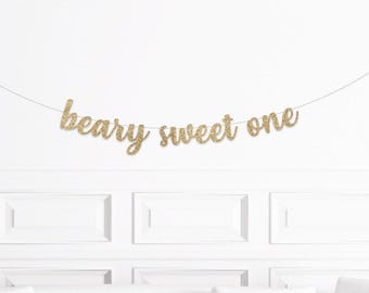May include: A gold glitter banner that reads "beary sweet one" in a cursive font. The banner is strung on a thin, silver string and is displayed against a white wall with decorative molding. This banner is perfect for a first birthday celebration.
