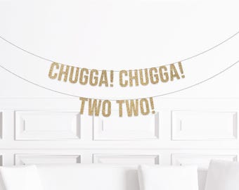May include: Gold glitter banner with the words "CHUGGA! CHUGGA!" and "TWO TWO!" strung across a white wall. The banner is a festive decoration, likely for a birthday celebration.