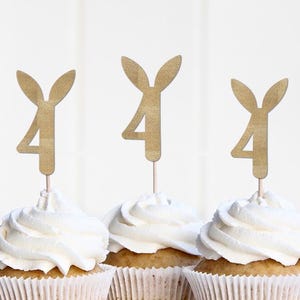 May include: Three cupcakes with white icing and gold-coloured bunny ear and number 4 toppers. The cupcakes are in white paper cases, ideal for a birthday party.