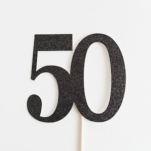 May include: A black glittery number 50 cake topper on a white background. The numbers are in a bold, sans-serif font and are attached to a wooden stick. The glitter adds a festive touch, perfect for a milestone birthday celebration.