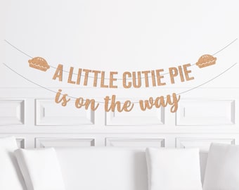 以下が含まれることがあります： 「A LITTLE CUTIE PIE is on the way」の文字が入ったゴールドグリッターのバナー。バナーには2つのパイのイラストが含まれています。白い壁と白いソファの上に吊るされています。