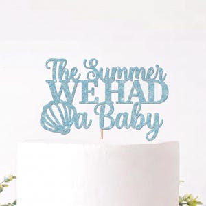 May include: A light blue glitter cake topper featuring the text The Summer WE HAD a Baby with a seashell graphic on the left side. The topper is attached to a thin wooden stick and is positioned above a plain white cake surface.