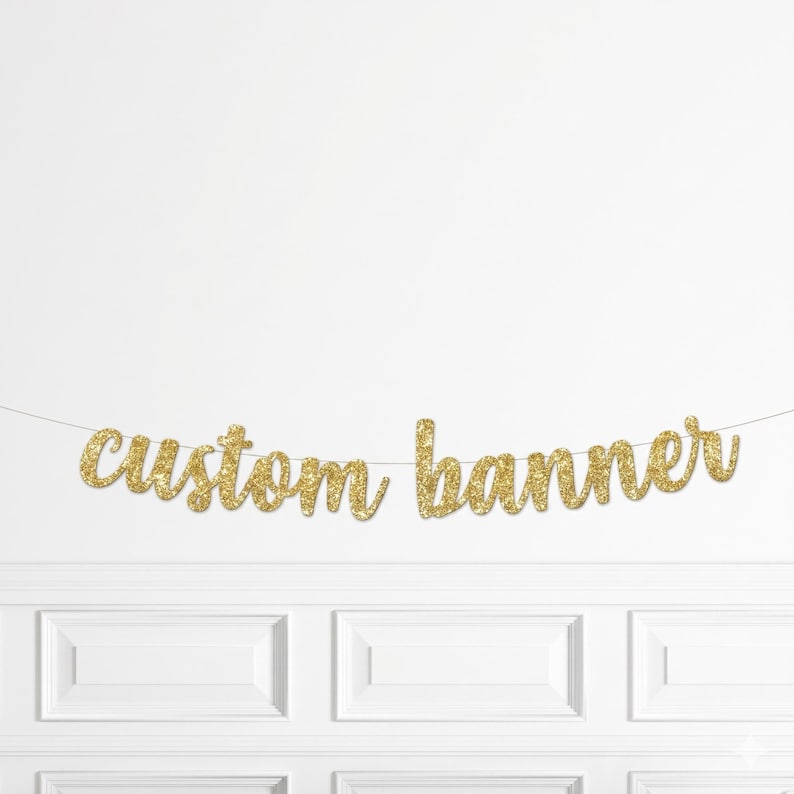Pu&ograve; includere: Un banner personalizzato con glitter dorati appeso a una parete bianca. Il banner &egrave; composto da singole lettere in corsivo. La parete ha pannelli bianchi sottostanti.