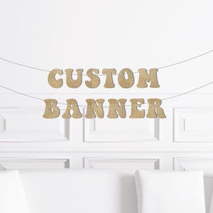 May include: A custom banner with the words "CUSTOM BANNER" in gold glitter letters strung across a thin black string. The banner hangs against a white wall with decorative molding and a white sofa.