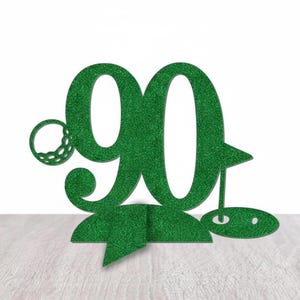 以下が含まれることがあります： 緑色のキラキラした「90」のケーキトッパー。ゴルフをテーマにしたデザインで、ゴルフボール、旗、グリーンが描かれています。ゴルフ好きの90歳の誕生日のお祝いに最適です。トッパーは自立するように設計されています。