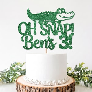 May include: A green glitter cake topper featuring a cartoon alligator and the text "OH SNAP! Ben's 3!" positioned on top of a white frosted cake. The topper is made of a sparkling material and is supported by a thin wooden stick.