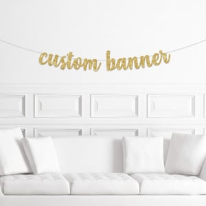 May include: A gold glitter "custom banner" hangs against a white wall above a white sofa with white cushions. The banner is strung on a thin, clear string. The text is in a cursive font.