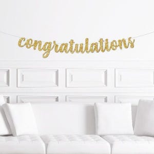 May include: A gold glitter "congratulations" banner is strung across a white wall. The banner is above a white couch with white pillows. The wall has a decorative panel design.