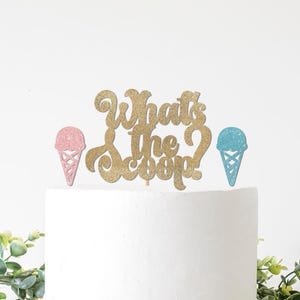 May include: A gold glitter cake topper with the words "What's the Scoop?" and two ice cream cone cutouts, one pink and one blue. The topper is on a white cake, with green foliage in the foreground.