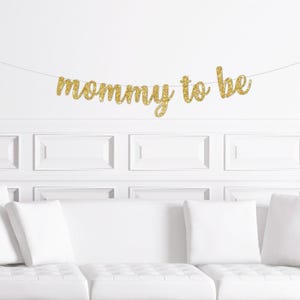 May include: A gold glitter banner reading "mommy to be" hangs against a white wall. Beneath the banner is a white sofa with white cushions. The banner is a decorative item for a baby shower or pregnancy announcement.