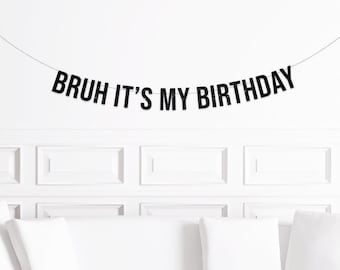 Pode incluir: Uma faixa preta com o texto "BRUH IT'S MY BIRTHDAY" está pendurada em uma parede branca. A faixa é presa por um fio preto fino. Abaixo da faixa, há uma parede branca com painéis retangulares e almofadas brancas.