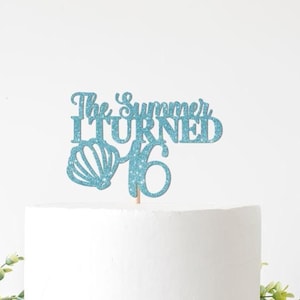 May include: A blue glitter cake topper with the text "The Summer I Turned 16" and a seashell design. The topper is on a white cake, with greenery at the base.