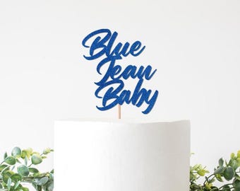 以下が含まれることがあります： 青いグリッターケーキトッパー。筆記体で「Blue Jean Baby」と書かれています。トッパーは木の棒に取り付けられ、白いケーキの上に置かれています。ケーキの底には緑の葉が見えます。