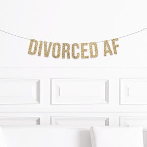 May include: A gold glitter banner that reads "DIVORCED AF" hangs against a white wall. The banner is strung on a thin black string. The wall has white paneling and a white couch is visible at the bottom.