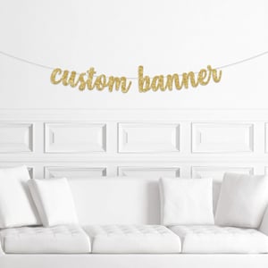 May include: A gold glitter banner that reads "custom banner" is strung across a white wall. The banner is in a cursive font. Below the banner is a white couch with white pillows.