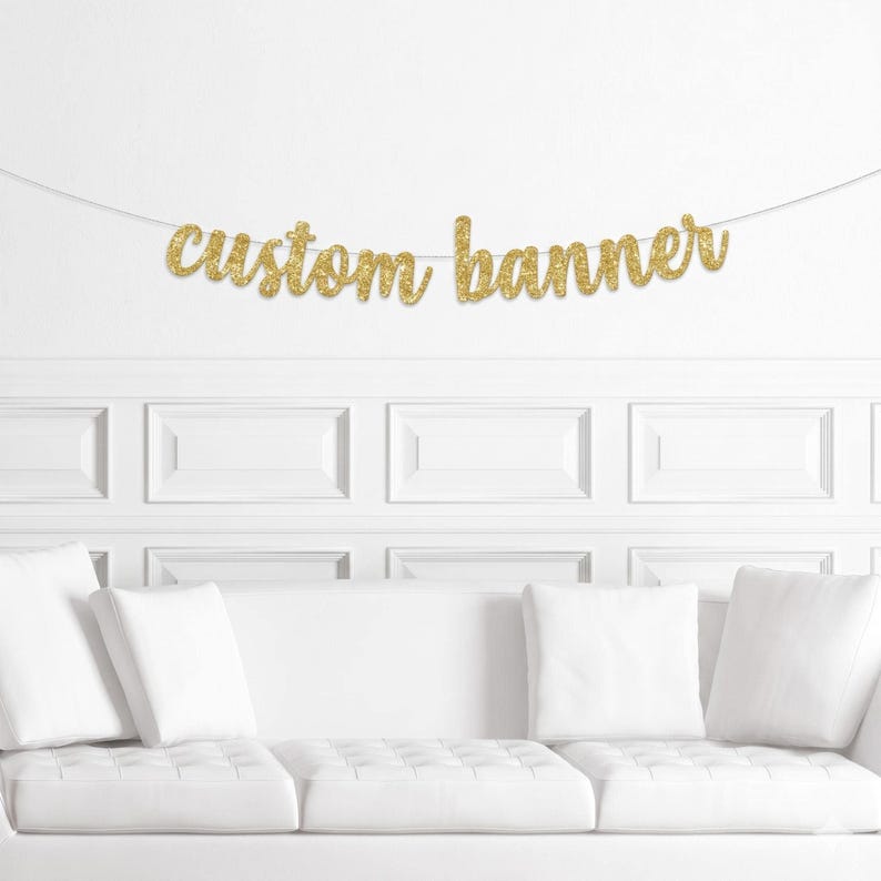 May include: A gold glitter banner that reads "custom banner" is strung across a white wall. The banner is in a cursive font. Below the banner is a white couch with white pillows.