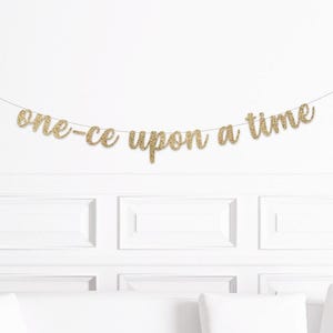 May include: A gold glitter banner that reads "one-ce upon a time" is strung across a white wall. The banner is suspended above a white couch and decorative wall panels.
