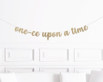 May include: A gold glitter banner that reads "one-ce upon a time" is strung across a white wall. The banner is suspended above a white couch and decorative wall panels.
