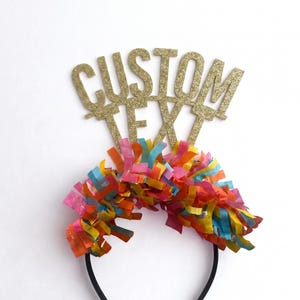 May include: A black headband featuring a gold glitter topper with the words CUSTOM TEXT in capital letters. The base of the topper is decorated with a dense fringe of multicolored paper strips in shades of pink, orange, yellow, and blue.
