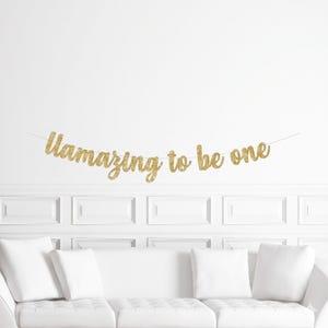 May include: A gold glitter banner that reads "Uamazing to be one" is strung across a white wall. The banner is above a white couch with white pillows. The banner is a festive decoration for a first birthday celebration.