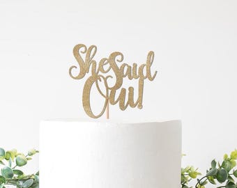 May include: A gold glitter cake topper that reads "She Said Oui!" in elegant script. The topper is attached to a wooden stick and is placed on top of a white cake. Greenery is visible in the background.