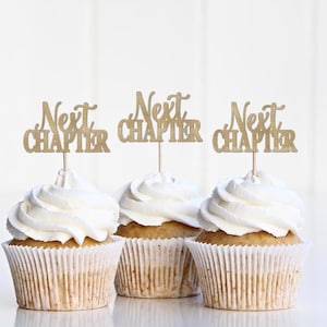 May include: Three cupcakes with white frosting and gold "Next Chapter" toppers. The cupcakes are in white paper liners and sit on a white surface. The text is in a cursive font.