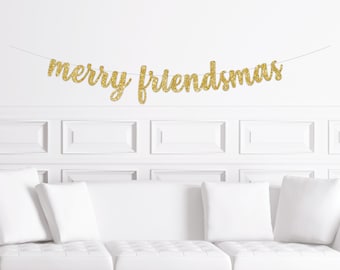 May include: Gold glitter banner with the text "merry friendsmas" hanging on a white wall above a white couch with white pillows.