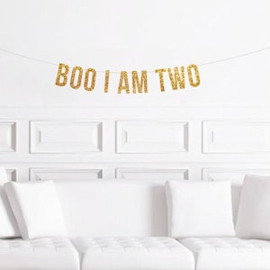 May include: A gold glitter banner that reads "BOO I AM TWO" hangs above a white couch with white pillows. The banner is strung across a white wall with decorative paneling. The banner is a festive decoration for a second birthday.