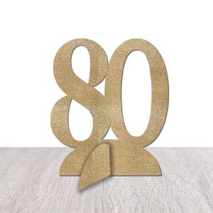 May include: A gold glitter-covered number 80, standing upright on a wooden surface. The number is cut from a solid material and has a stand attached to the bottom for stability. The background is white.