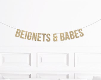 May include: A gold glitter banner strung across a white wall. The banner reads "BEIGNETS & BABES" in capital letters. The letters are made of gold glitter and are strung on a thin black string.
