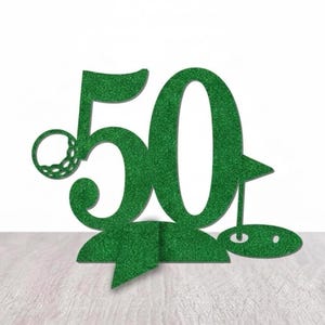 May include: A green glittery golf-themed 50th birthday cake topper. The design features the number 50 with a golf ball and a golf flag. The topper is designed to stand upright on a flat surface.