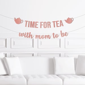 May include: A rose gold glitter banner reading "TIME FOR TEA with mom to be" hangs against a white wall. The banner features two teapot illustrations. A white sofa with white cushions is in the foreground.