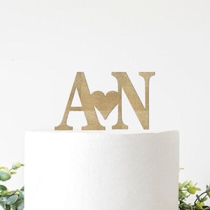 May include: A gold-colored wooden cake topper featuring the letters "A" and "N" with a heart in between. The topper is placed on top of a white cake, with greenery in the background. A wedding or anniversary theme.